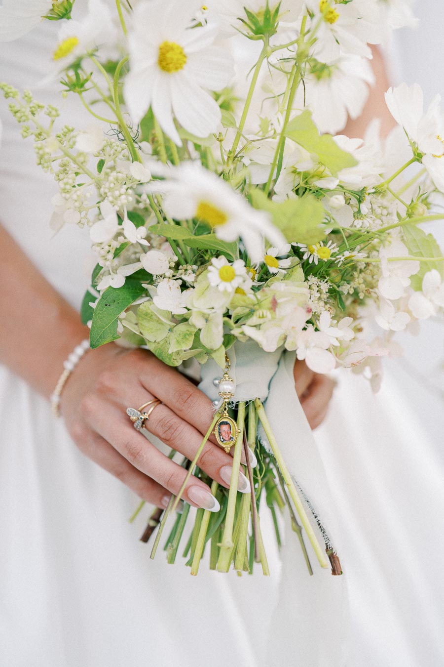 Bridal hand holding a bouquet of white daisies and greenery, adorned with a locket pendant, showcasing wedding elegance and delicate floral arrangement.