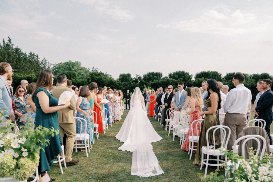 Outdoor wedding ceremony with a bride walking down the aisle, surrounded by standing guests dressed in colorful attire, set against a backdrop of lush greenery and a clear sky.