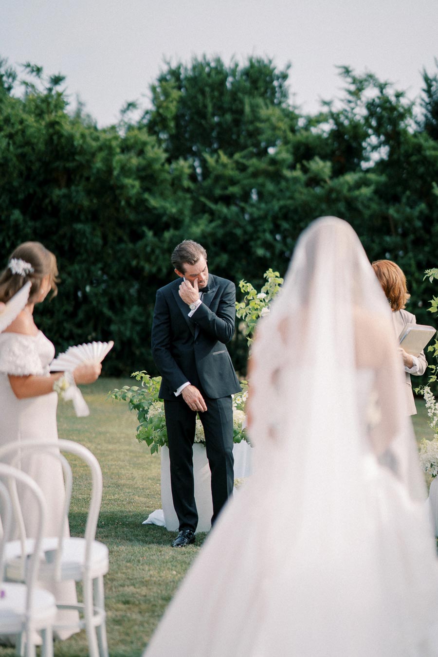 Emotional groom in black suit wiping tears during outdoor wedding ceremony, bride in foreground with veil, surrounded by lush greenery and guests.