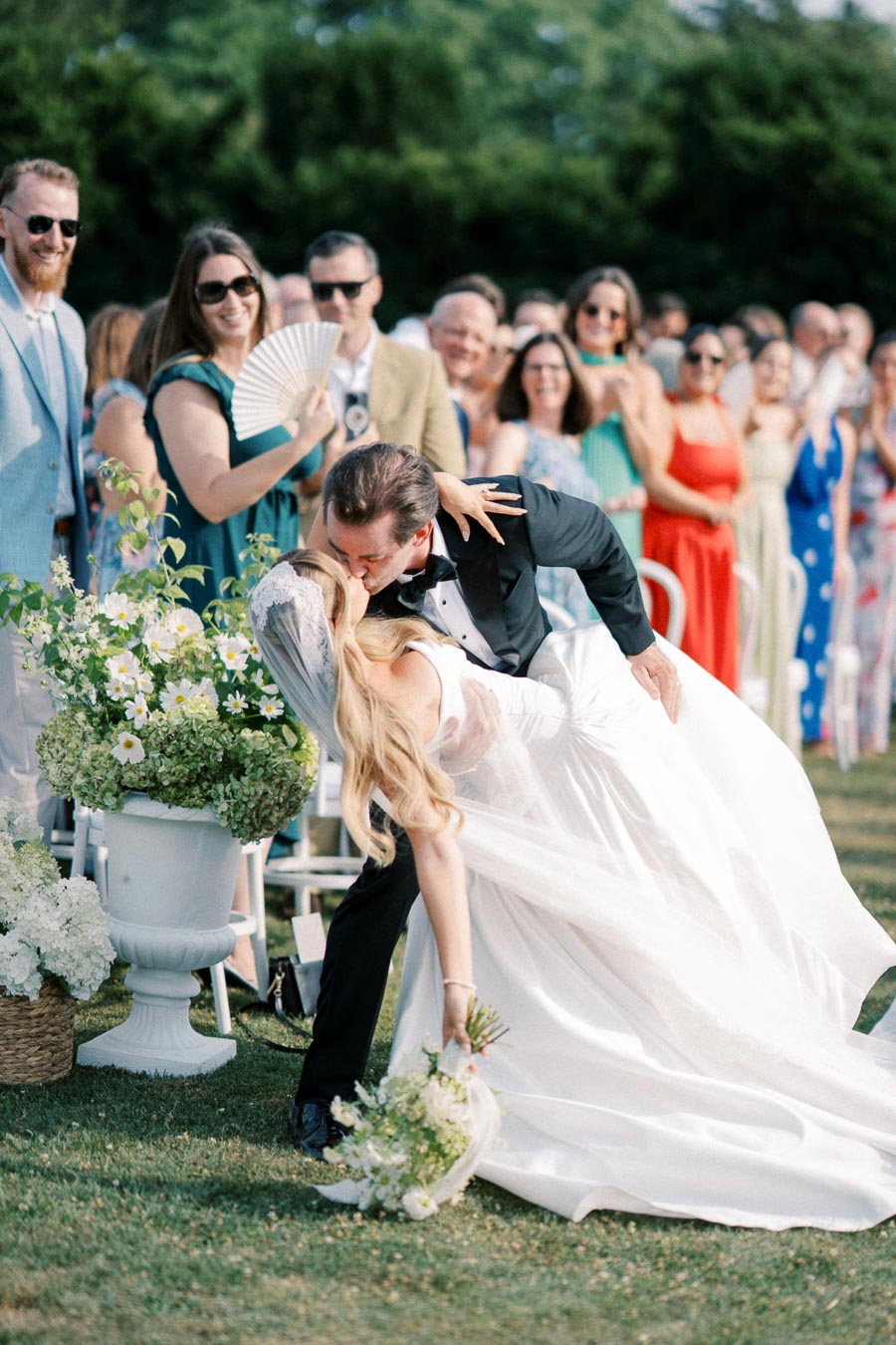 A newlywed couple shares a romantic kiss in front of joyful wedding guests, with the bride in a flowing white gown and veil and the groom in a black tuxedo, surrounded by lush garden flowers.