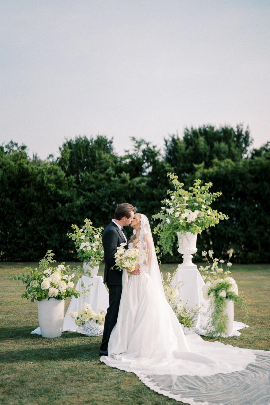 Outdoor wedding ceremony with bride and groom kissing, surrounded by lush greenery and floral arrangements, elegant white dress and suit, romantic garden setting