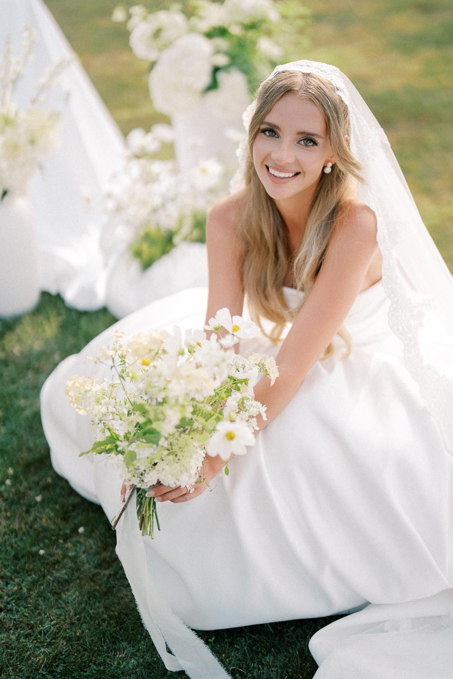 A bride in a white wedding dress and veil smiling while holding a bouquet of white and green flowers, sitting outdoors on a sunny day.