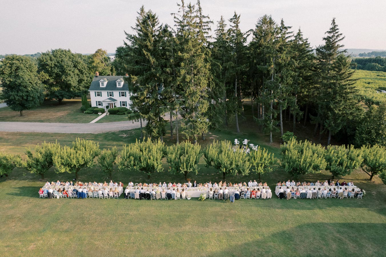 Aerial view of an outdoor banquet event with long tables set on a lush green lawn, surrounded by trees, near a classic white farmhouse and a small forest in the background.