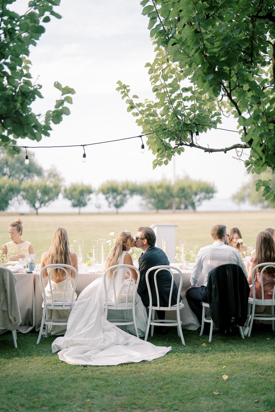 Outdoor wedding reception with a bride and groom kissing at a long table under string lights, surrounded by guests, with a scenic landscape in the background.