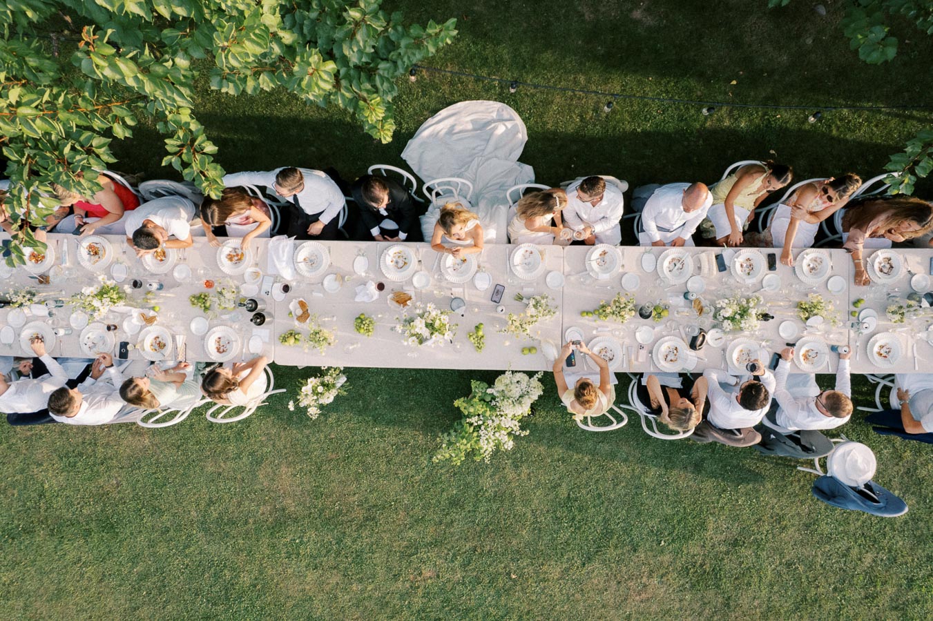 Aerial view of an elegant outdoor dining setup, featuring a long table adorned with white tablecloths, floral centerpieces, and neatly arranged place settings. People dressed in formal attire are gathered around the table, enjoying a meal in a lush garden setting, surrounded by greenery.