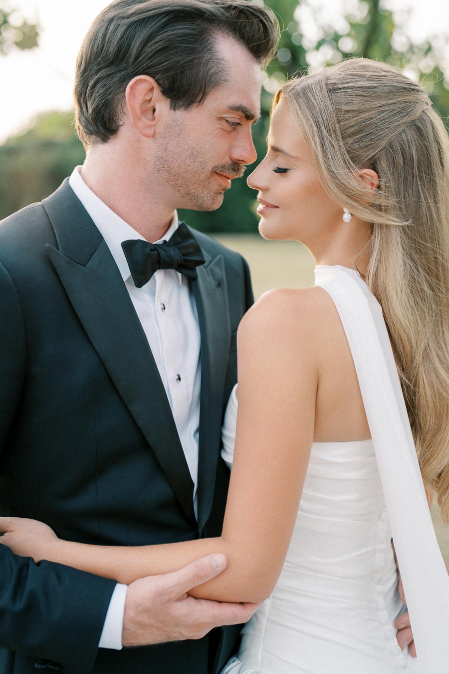 Alt text: Elegant bride and groom embracing outdoors, groom in black tuxedo and bride in white gown, with natural greenery background.