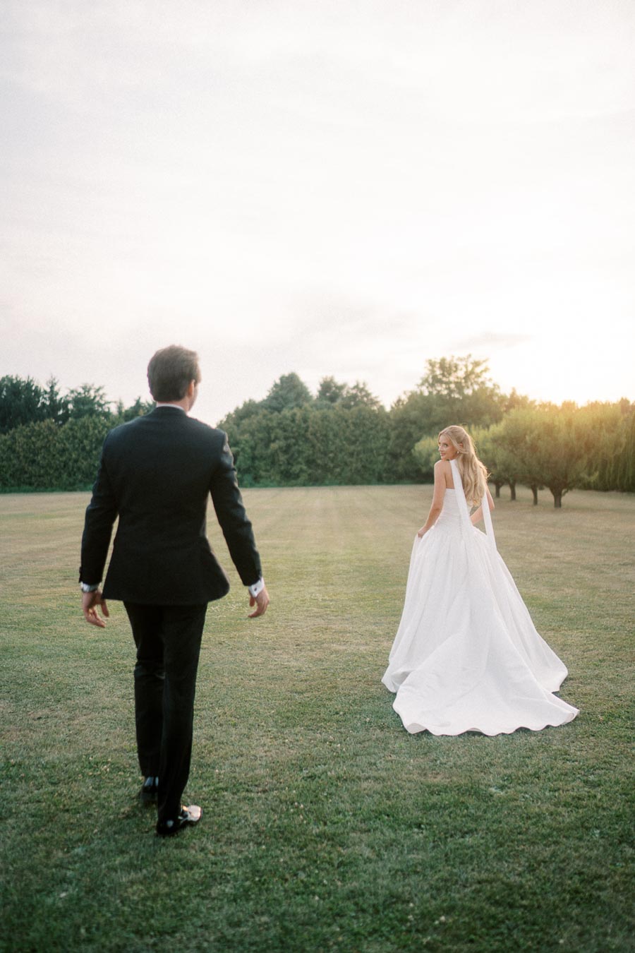 A bride in a flowing white gown and a groom in a black suit walking together on a grassy field during sunset, surrounded by lush greenery.