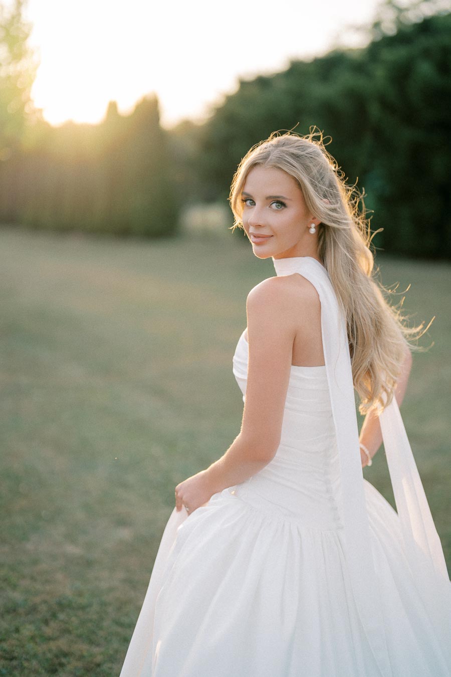 Blonde woman in a flowing white wedding dress standing outside at sunset, with scenic greenery in the background.