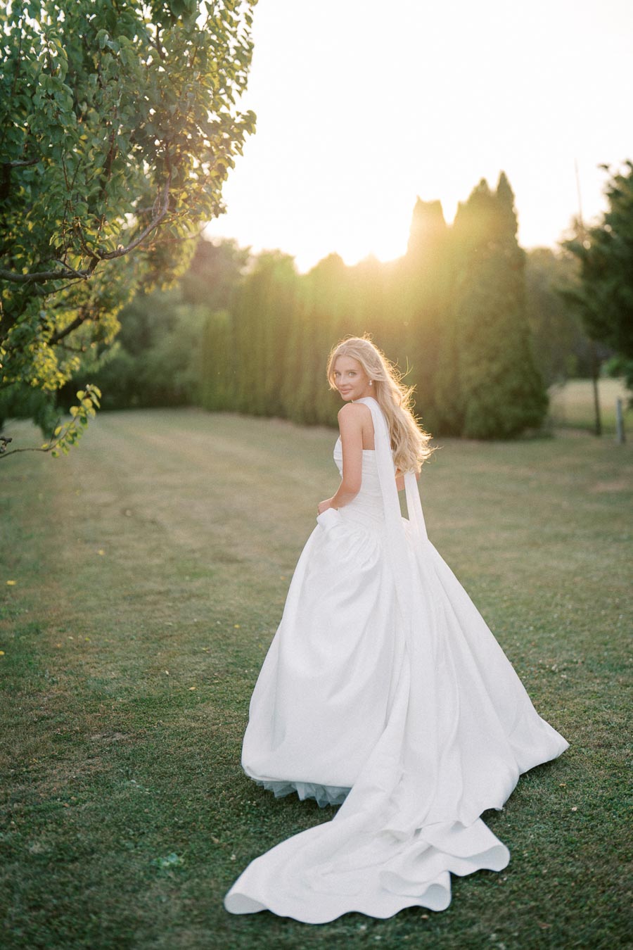 A bride in a flowing white wedding dress walks through a sunlit garden, with long grass and trees in the background, creating a serene and romantic setting.