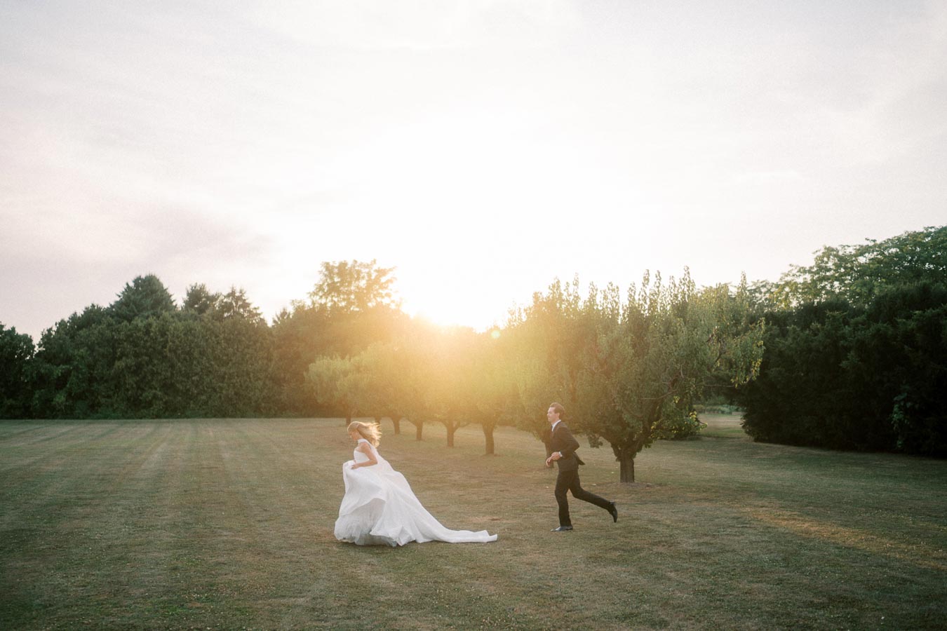 A bride and groom joyfully run across a sunlit field surrounded by lush green trees, capturing a romantic wedding moment at sunset.