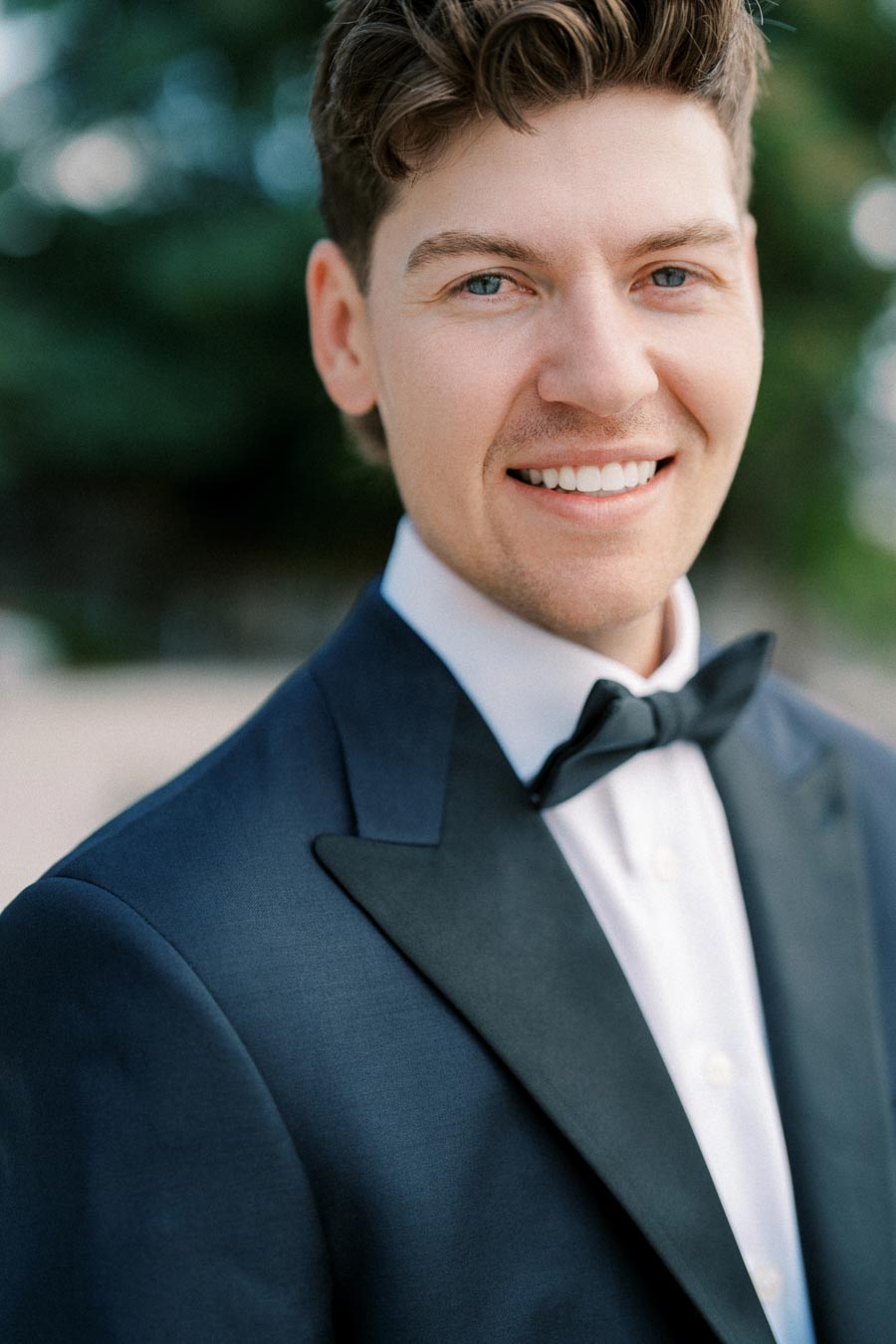 ```markdown
Alt text: Smiling man in formal suit with bow tie, standing outdoors with blurred green background.
```