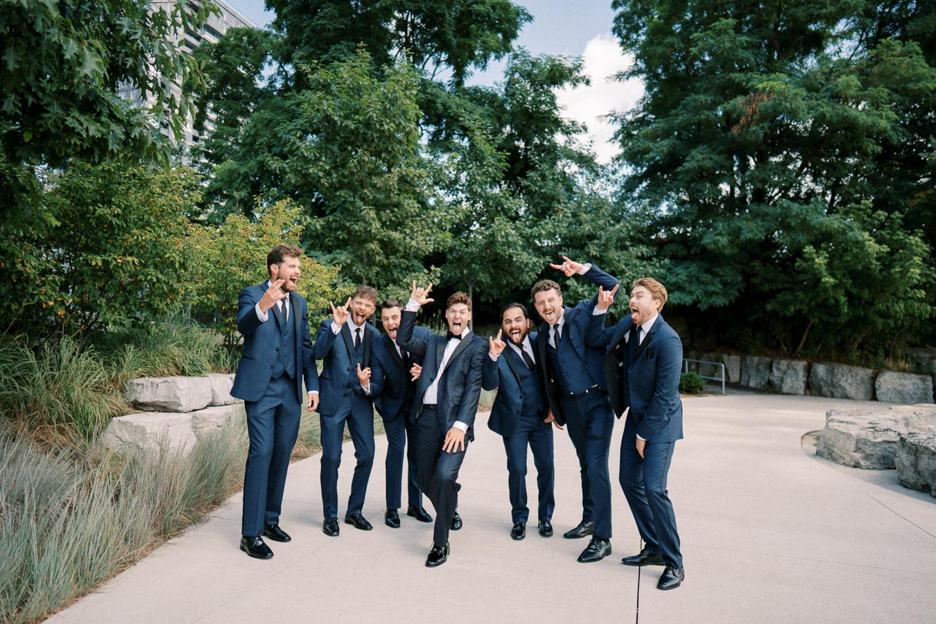 A group of groomsmen in blue suits making playful gestures and expressions outdoors with trees in the background, creating a fun and lively wedding moment.