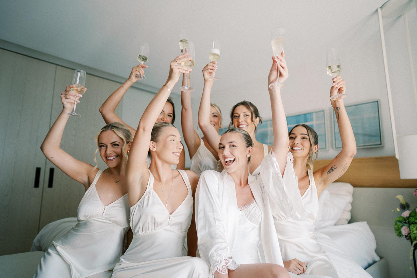 A group of smiling bridesmaids and a bride in white robes raising champagne glasses in a joyful celebration.