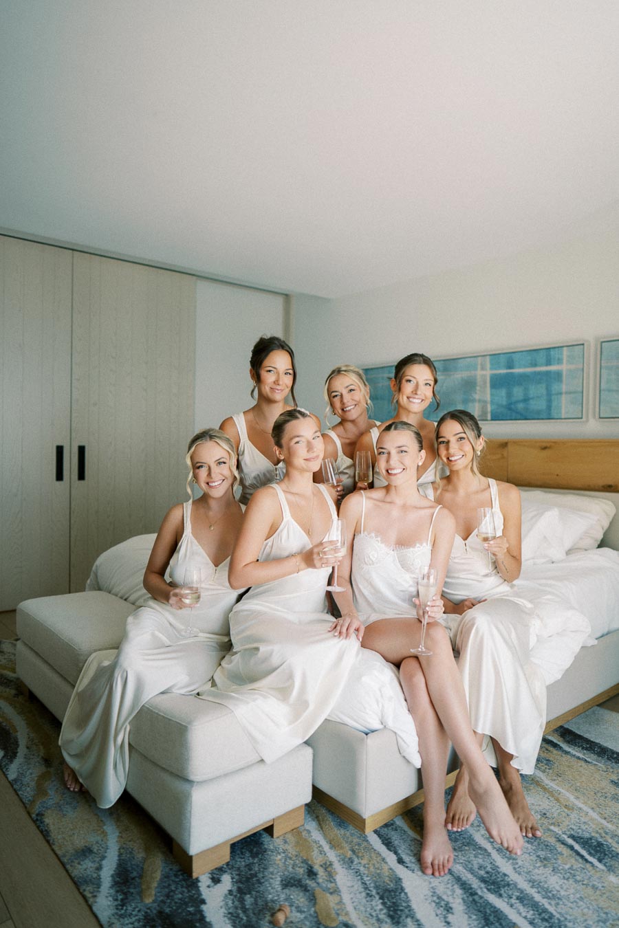 Bridesmaids in matching white robes smile and hold champagne glasses while sitting on a bed in a bright, elegant room, celebrating before a wedding.
