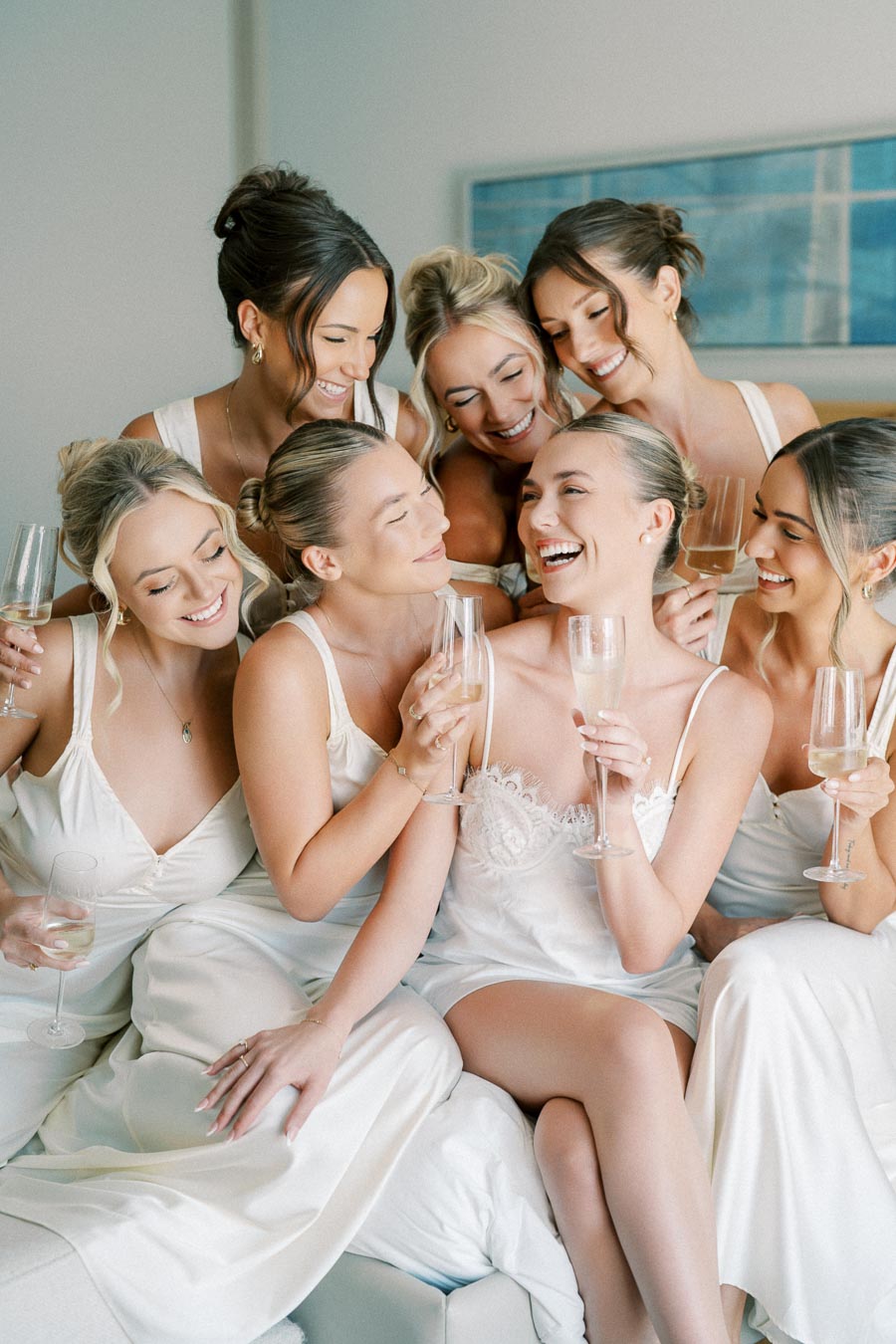 Bridal party laughing and toasting with champagne, wearing matching white robes, celebrating a wedding morning.