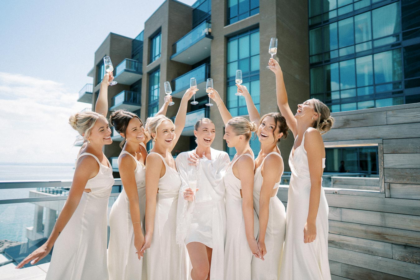 Bridesmaids in matching elegant white dresses celebrating with champagne on a hotel balcony, featuring modern architecture in the background.