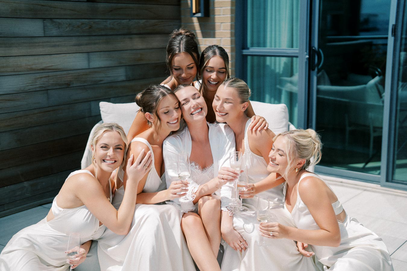 A joyful bridal party of seven women in white robes and dresses celebrating with champagne outdoors on a sunny day.