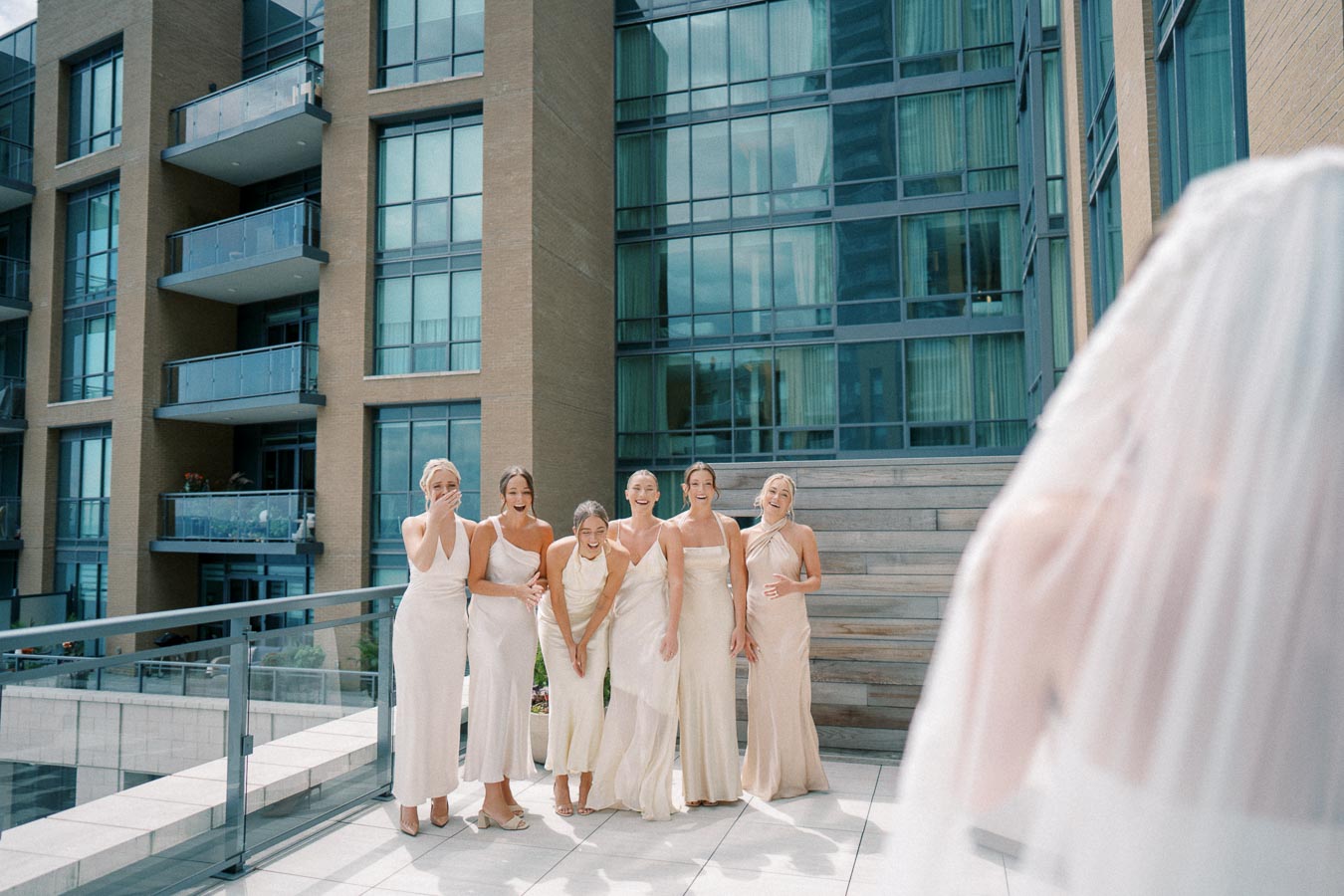 Bridesmaids dressed in elegant light beige dresses react joyfully on a modern urban rooftop as the bride approaches, showcasing a contemporary city wedding setting.