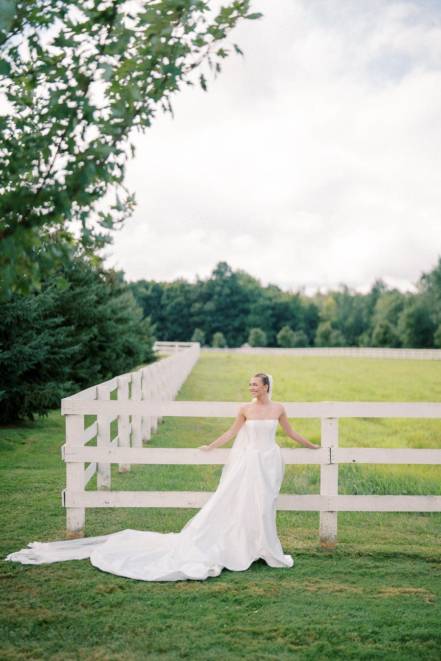Bride in a flowing white wedding dress standing by a white fence in a lush green countryside setting with trees in the background.
