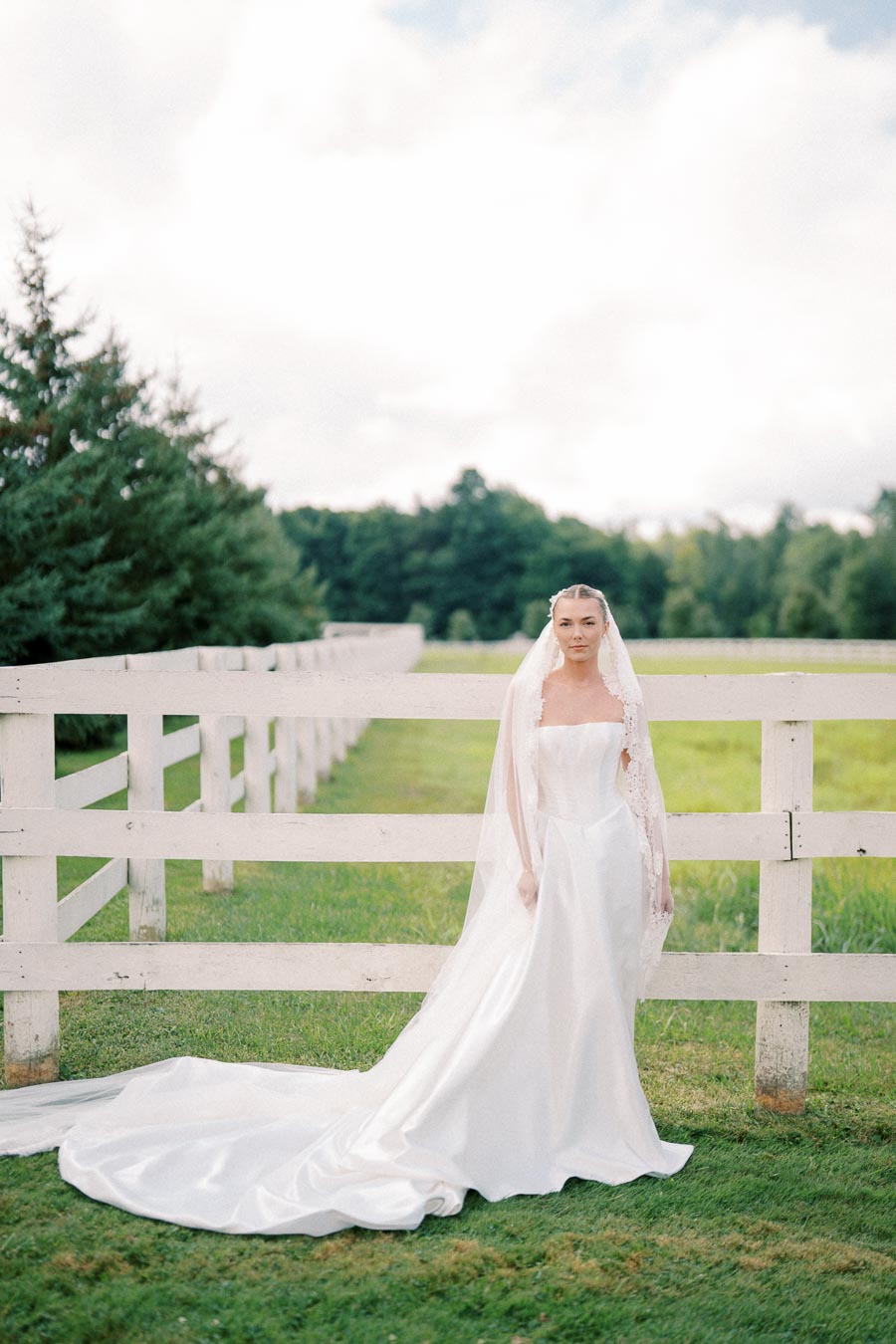 Bride in a flowing white wedding dress and veil standing by a rustic wooden fence in a lush green field, under a cloudy sky.