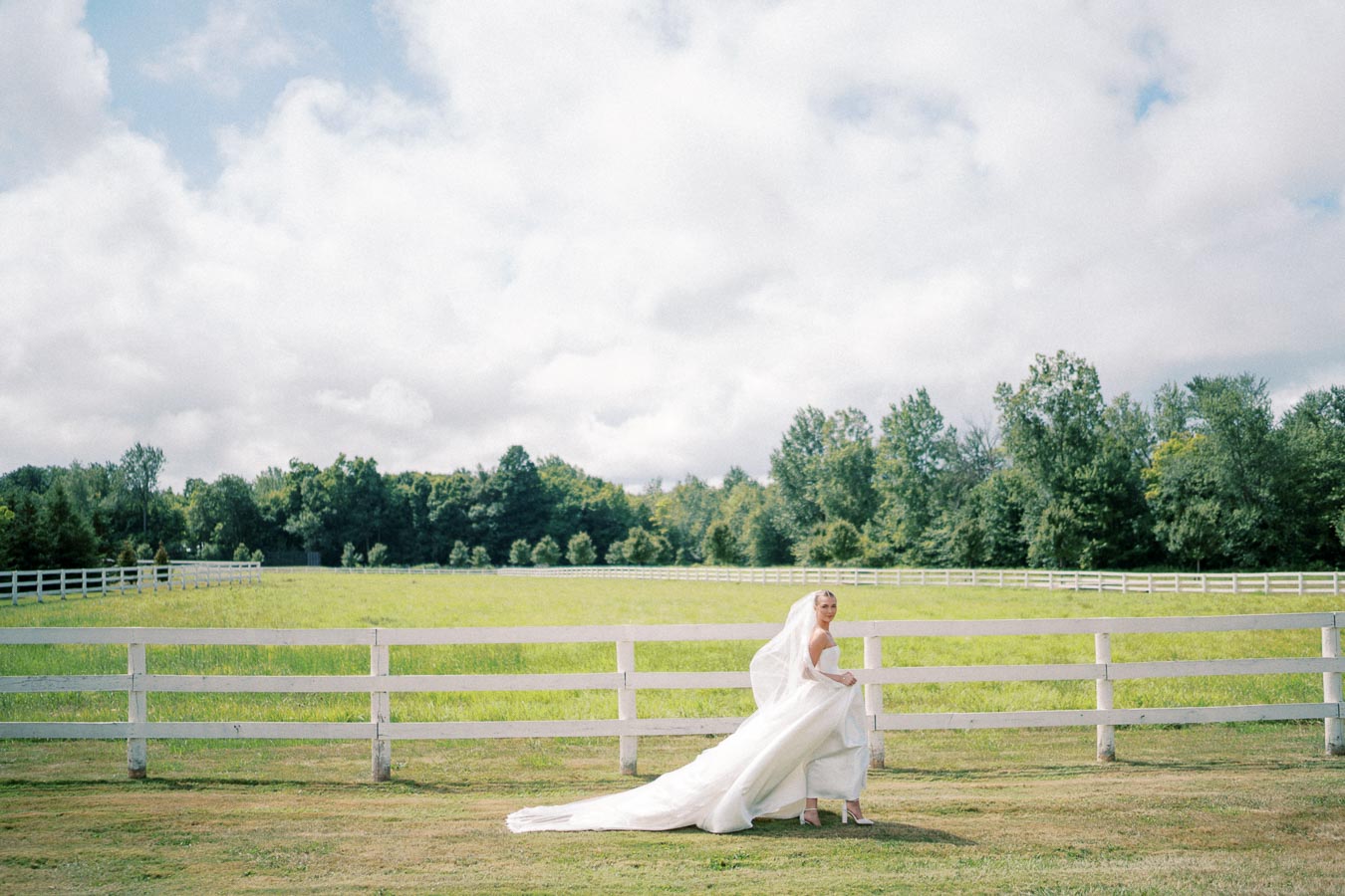 Bride in an elegant white wedding gown standing by a rustic fence in a picturesque green field, with a lush forest backdrop and a cloudy blue sky.