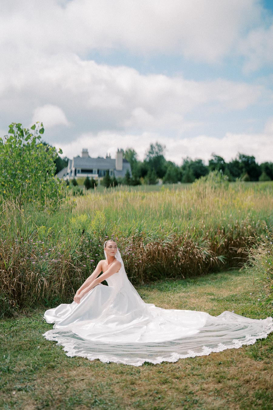 A bride in an elegant white wedding dress with a long train sits gracefully in a lush green field with a picturesque house and trees in the background under a partly cloudy sky.