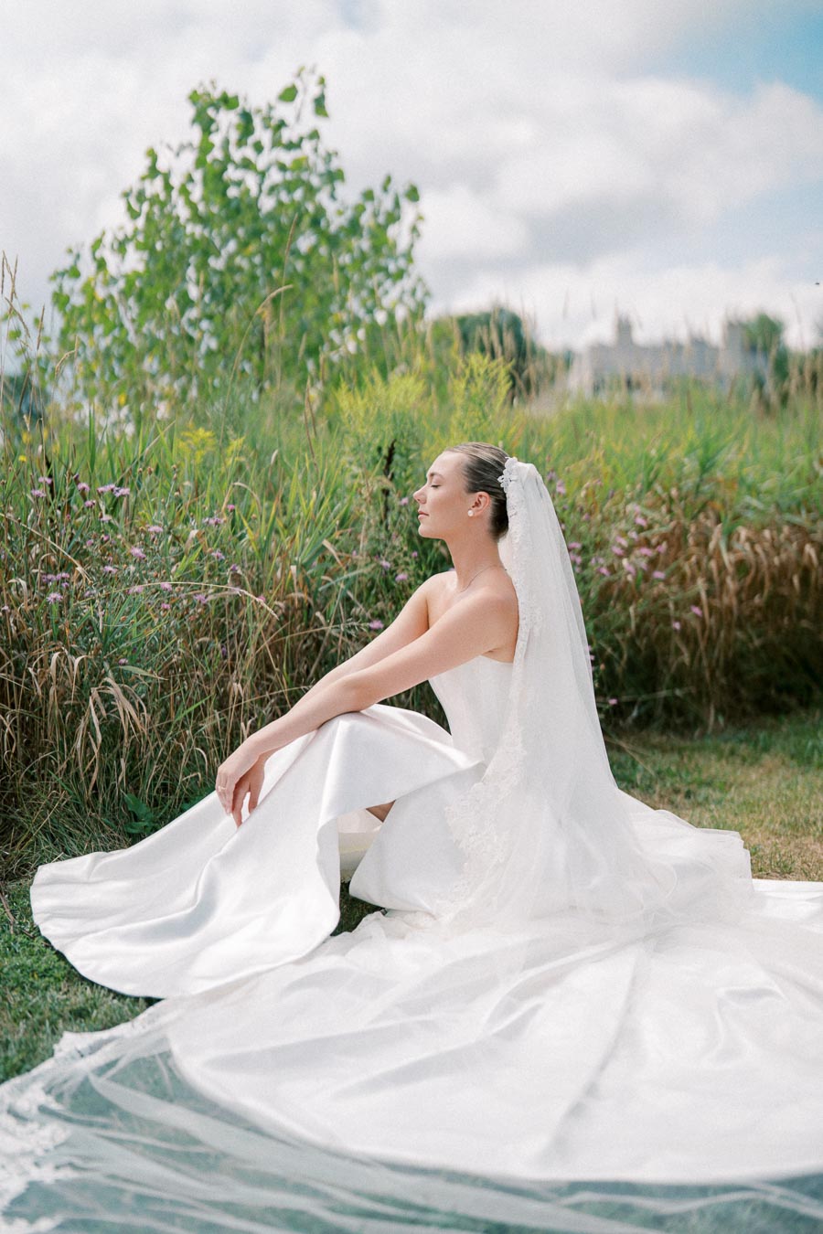 A bride in an elegant white wedding dress sits in a serene outdoor setting, surrounded by lush greenery and wildflowers, with a cloudy sky in the background.