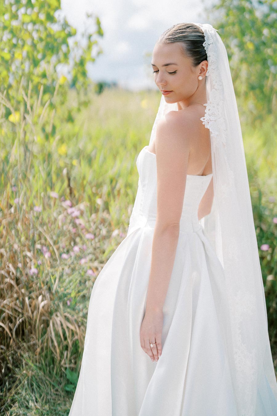A bride in an elegant white wedding dress and lace veil stands in a sunlit meadow, surrounded by greenery and wildflowers, with a serene expression on her face.
