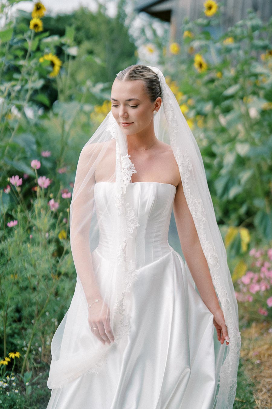 A bride in a strapless white wedding dress and lace veil stands outdoors, surrounded by lush green plants and blooming sunflowers.