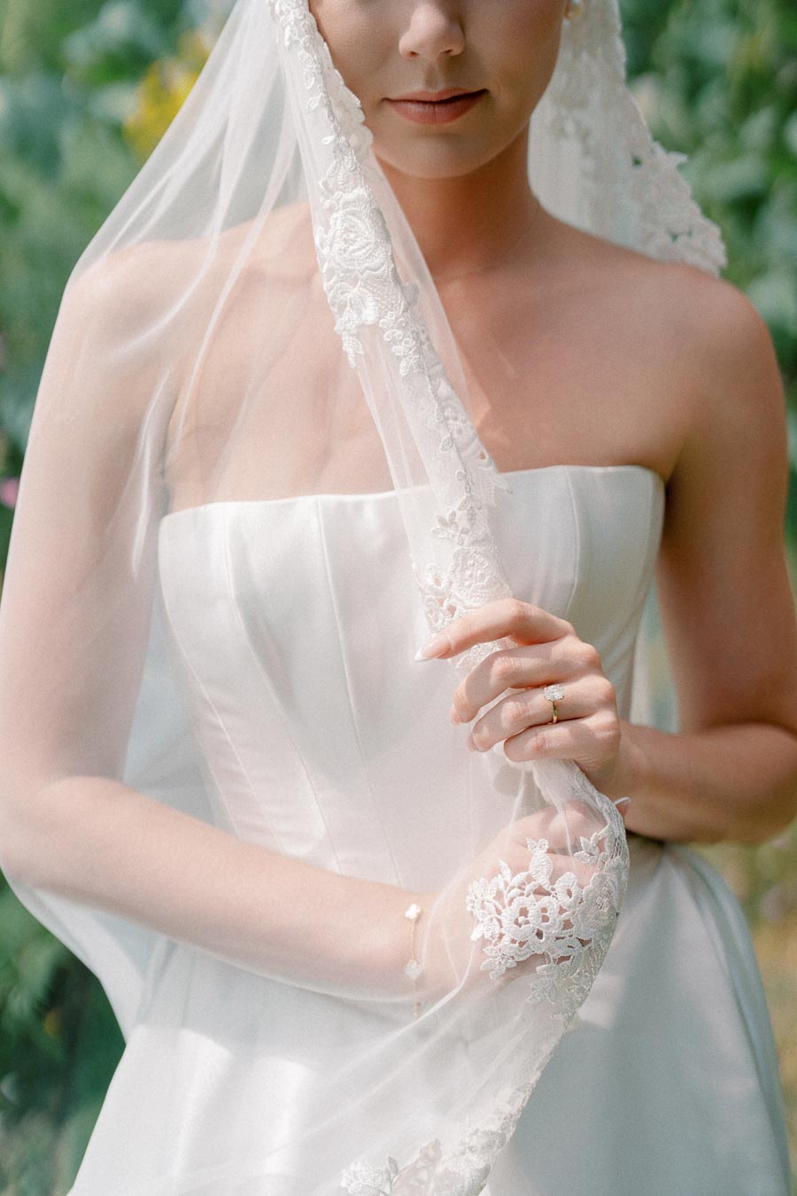 Elegant bride in a strapless wedding dress holding a delicate lace-edged veil.