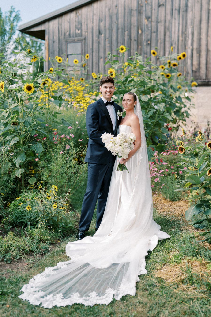 A bride in a flowing white gown and veil stands beside a groom in a classic black tuxedo, both surrounded by vibrant sunflowers and wildflowers, with a rustic wooden barn in the background.