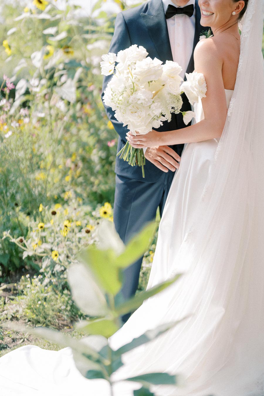 Bride and groom embracing in a garden, with the bride holding a bouquet of white flowers, and greenery in the background, capturing a romantic wedding moment.