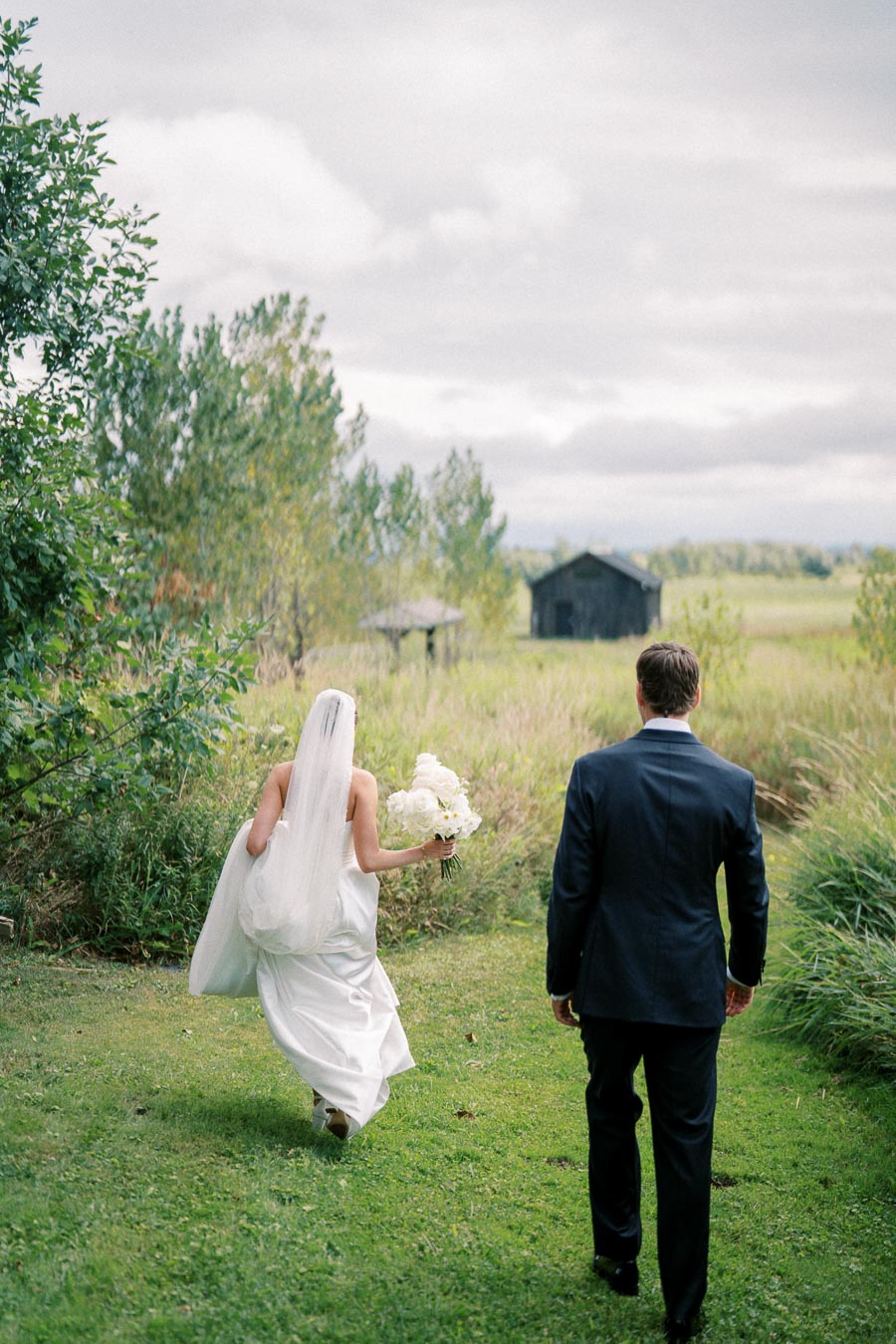 Bride and groom walking through lush greenery towards a rustic barn, with the bride in a white dress holding a bouquet of flowers.