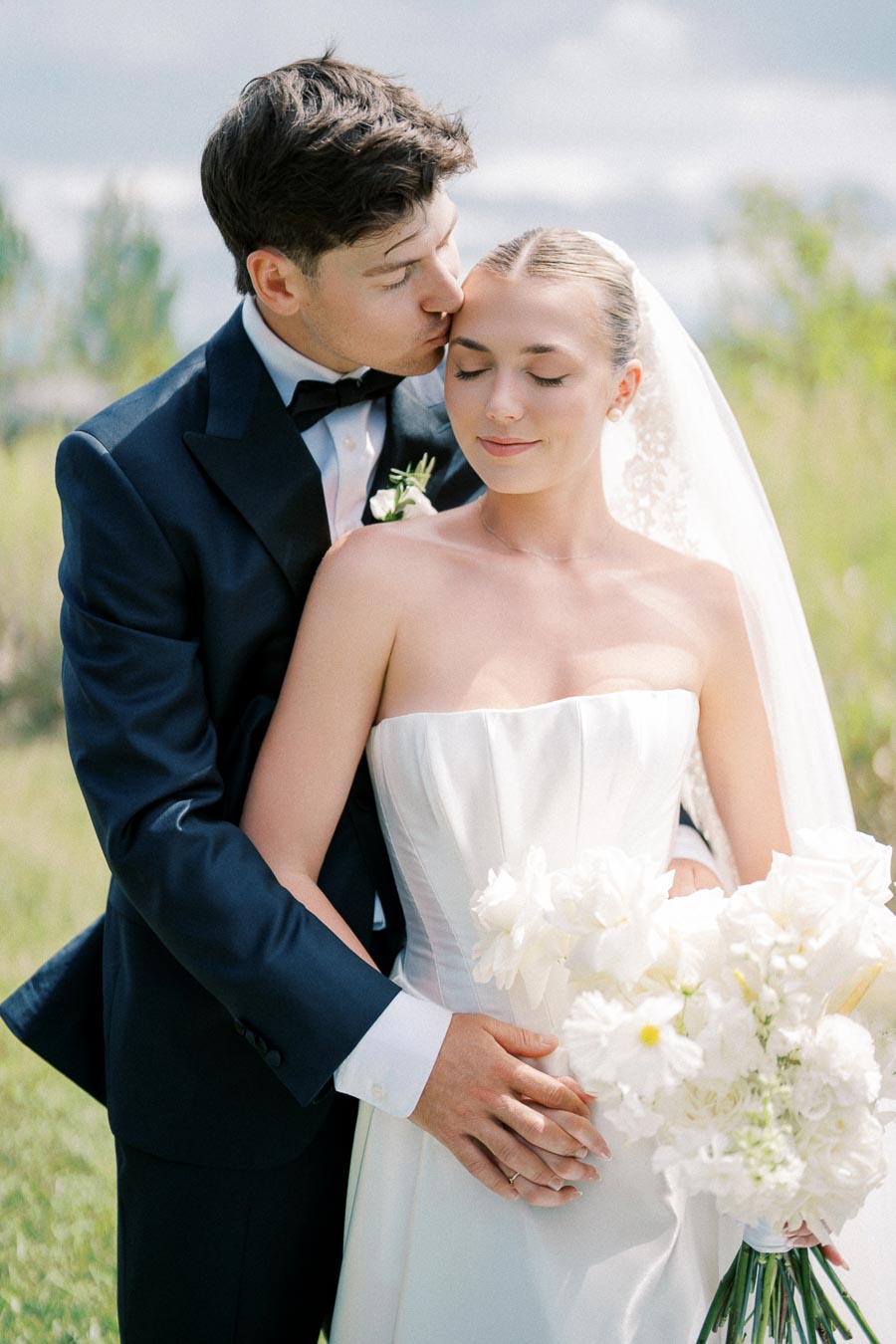 Elegant bride in strapless wedding dress holding white bouquet while groom in black suit gently kisses her forehead outdoors on a sunny day