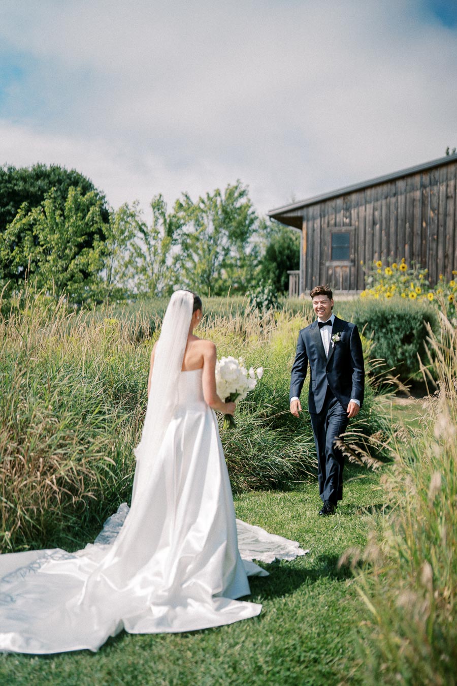 Bride in elegant white gown holding flowers, meets groom in dark suit outdoors by rustic wooden building with greenery and a clear sky.