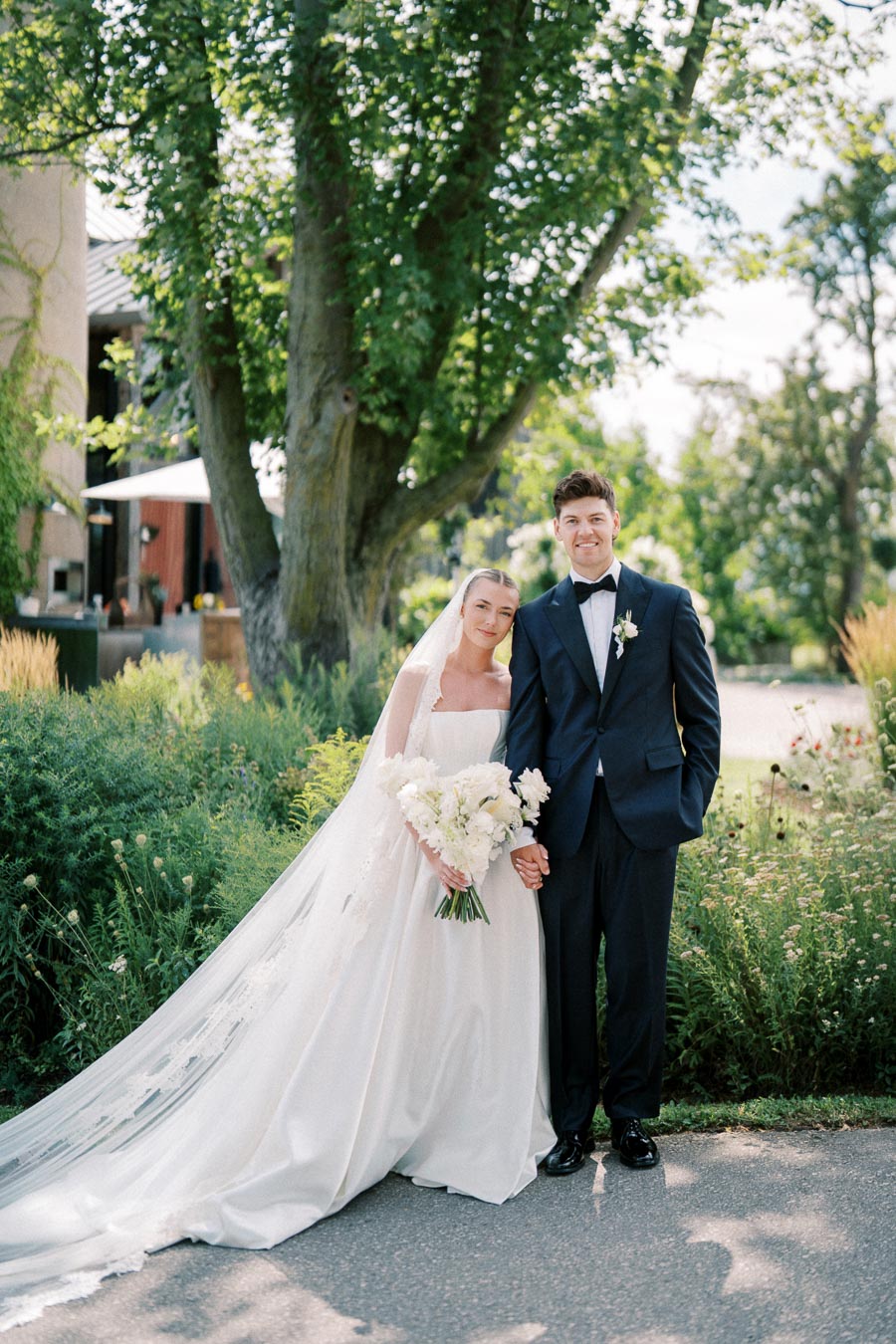 A bride and groom standing hand in hand, dressed in elegant wedding attire, pose outdoors amidst lush greenery and tall trees on a sunny day. The bride holds a bouquet of white flowers, and the groom wears a dark suit with a boutonniere.