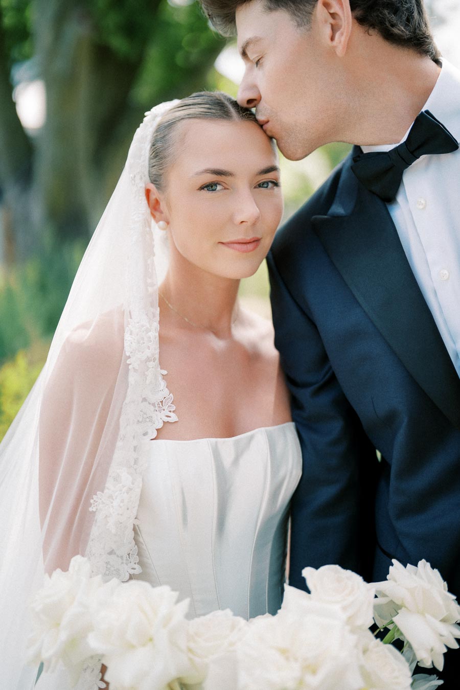 A bride in a white lace wedding dress and veil stands with a bouquet of white roses while being gently kissed on the forehead by a groom in a navy tuxedo, set outdoors against a soft-focus natural background, capturing an intimate and romantic moment.