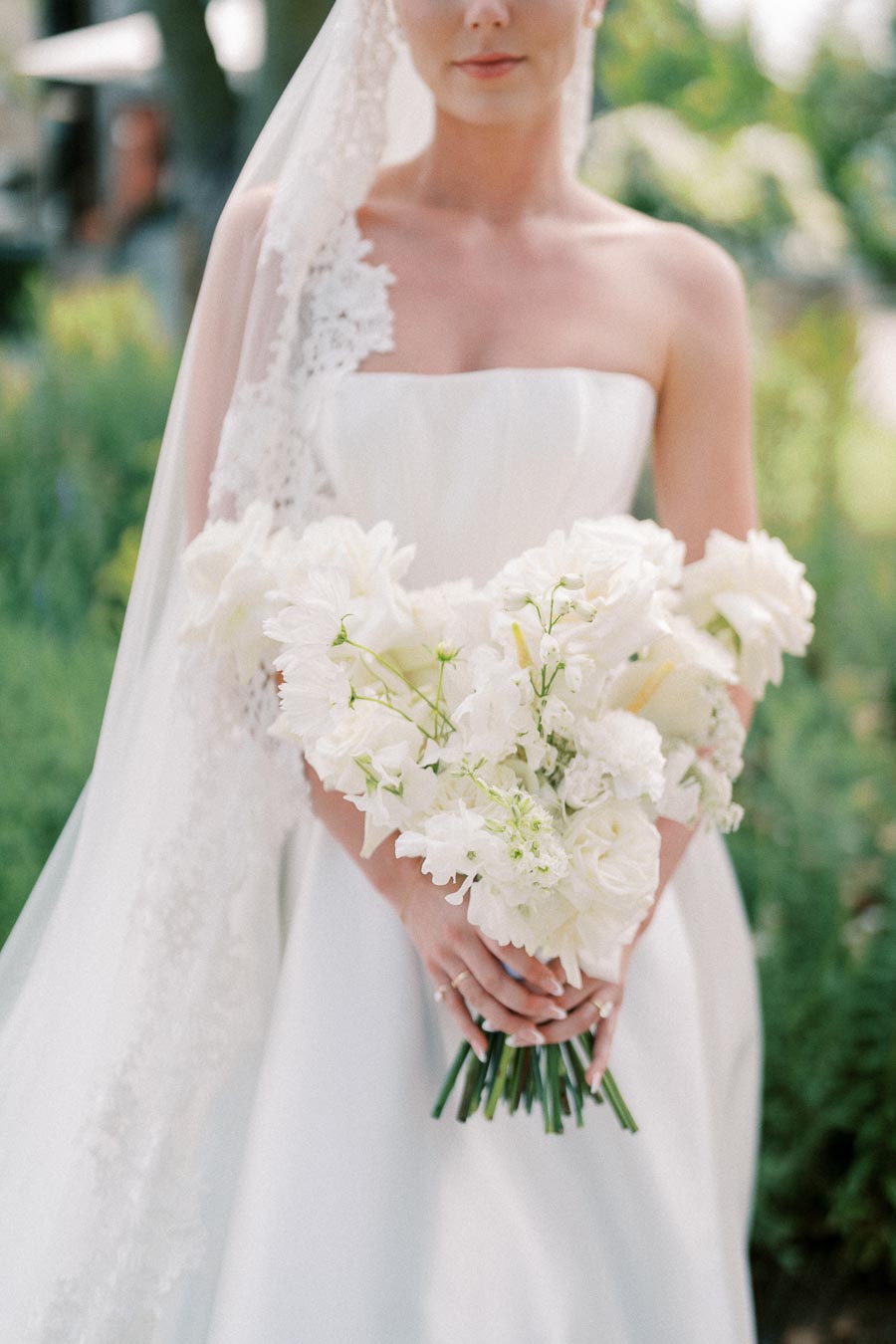 A bride in a white strapless wedding dress holds a bouquet of white flowers, with a lace-trimmed veil cascading from her head, set against a blurred outdoor background.