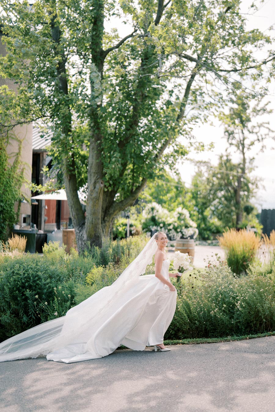 Bride smiling in an elegant white wedding dress with a long train, walking along a garden path with lush greenery and trees on a sunny day.