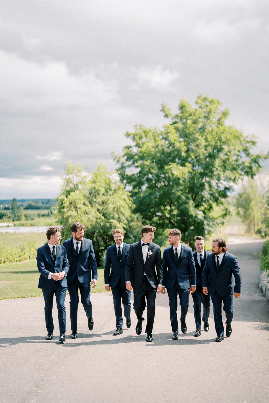 A group of seven men in matching navy suits and ties, walking outdoors on a sunny day, surrounded by lush green trees and a clear sky.