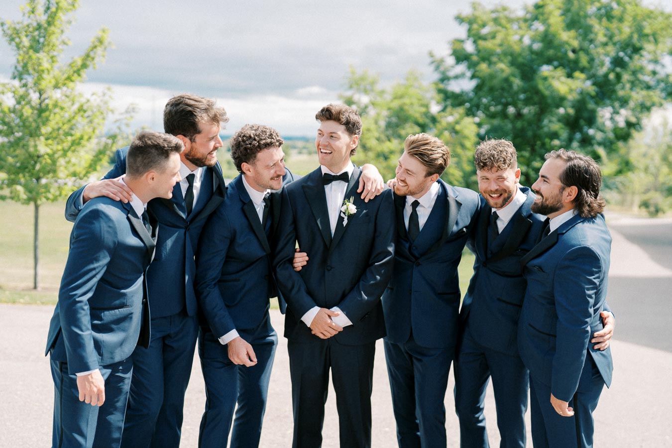 Groom and groomsmen in blue suits smiling and standing together outdoors, celebrating on a sunny day.