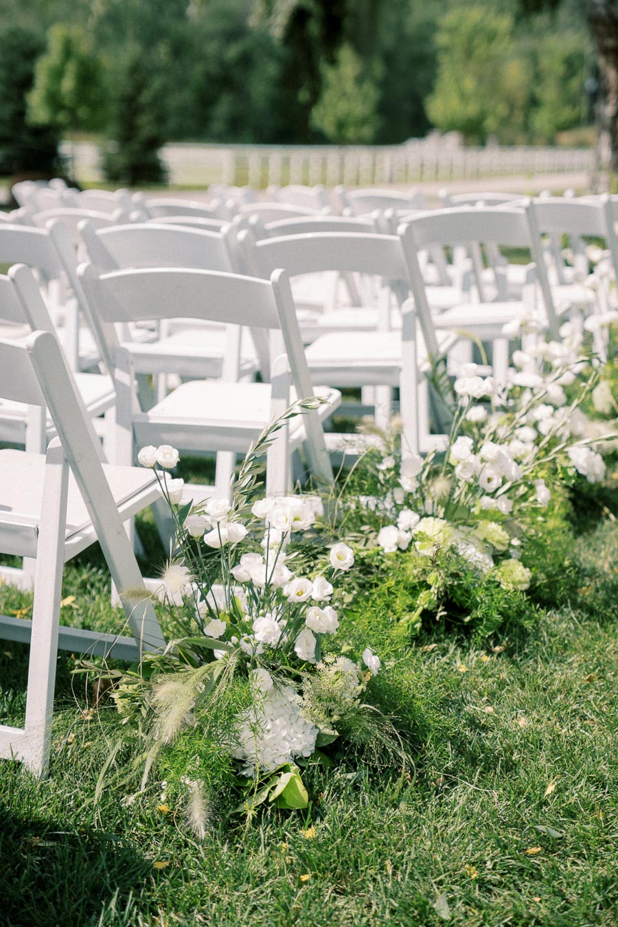 Rows of white chairs lined up on lush green grass, adorned with elegant white floral arrangements, set for an outdoor wedding ceremony.