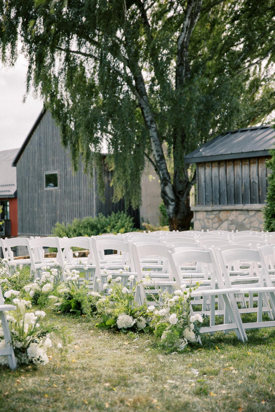 Outdoor wedding ceremony setup with rows of white chairs decorated with greenery and white flowers, set against a rustic barn backdrop and lush green trees.