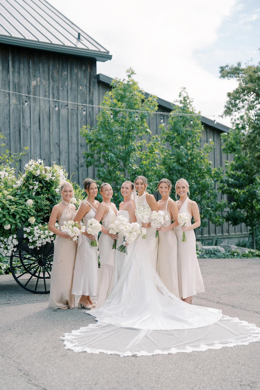 A bride in a white gown with a long train, surrounded by bridesmaids in matching pale dresses, holding white floral bouquets, posing outdoors in front of rustic wooden barn and lush greenery.