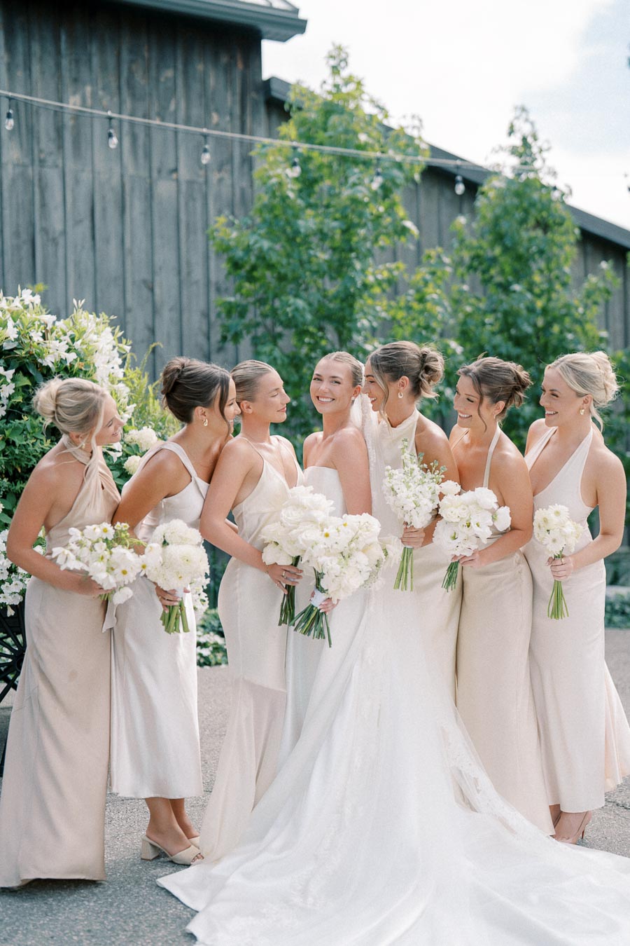 Group of bridesmaids in beige dresses with a bride in a white gown, holding white floral bouquets outdoors at a rustic wedding venue.