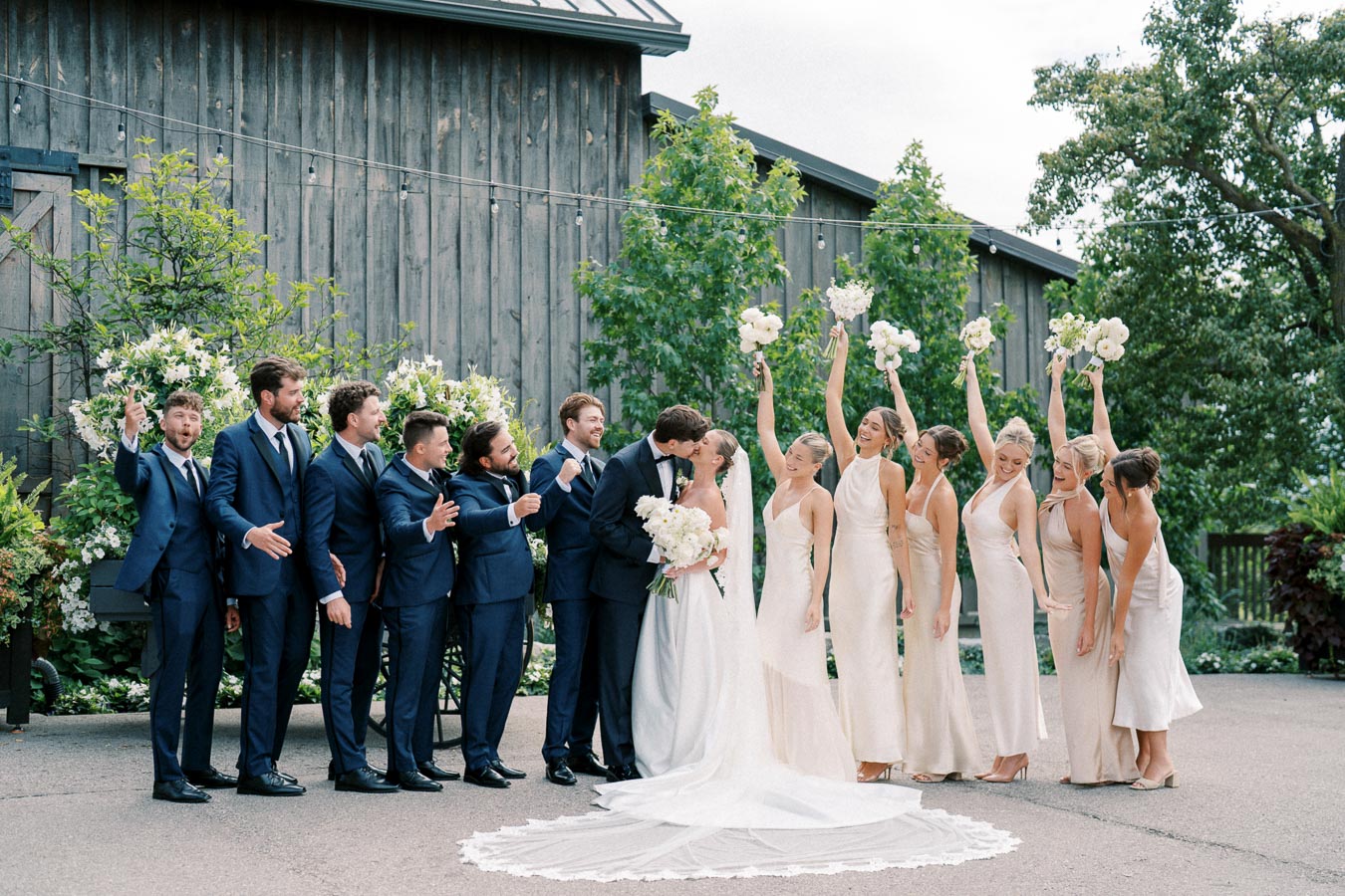 Wedding party posing outdoors, featuring bride and groom kissing, with bridesmaids in light dresses and groomsmen in navy suits, celebrating against a rustic barn backdrop.