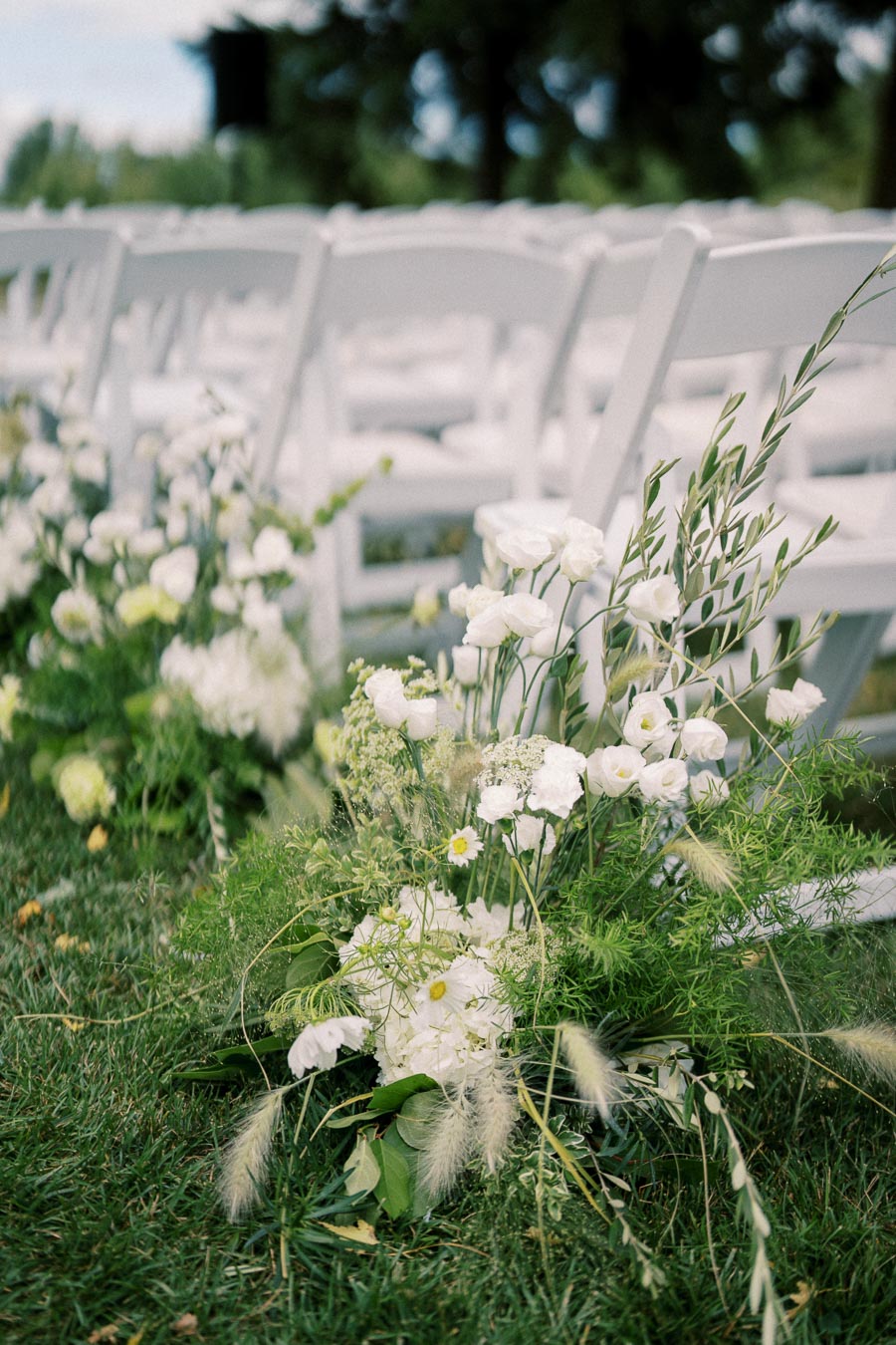 Ceremony aisle decorated with lush white flowers and greenery beside arranged white chairs outdoors, perfect for a garden wedding setting.
