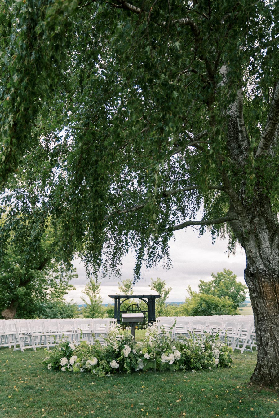 Outdoor wedding ceremony setup with white chairs arranged in rows, lush greenery, and floral arrangements under a large tree.