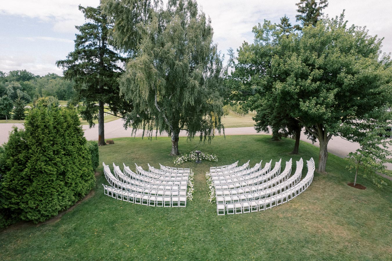 Aerial view of an outdoor wedding ceremony setup with white chairs arranged in a semi-circle on a lush green lawn, surrounded by large trees and floral decorations under a clear sky.
