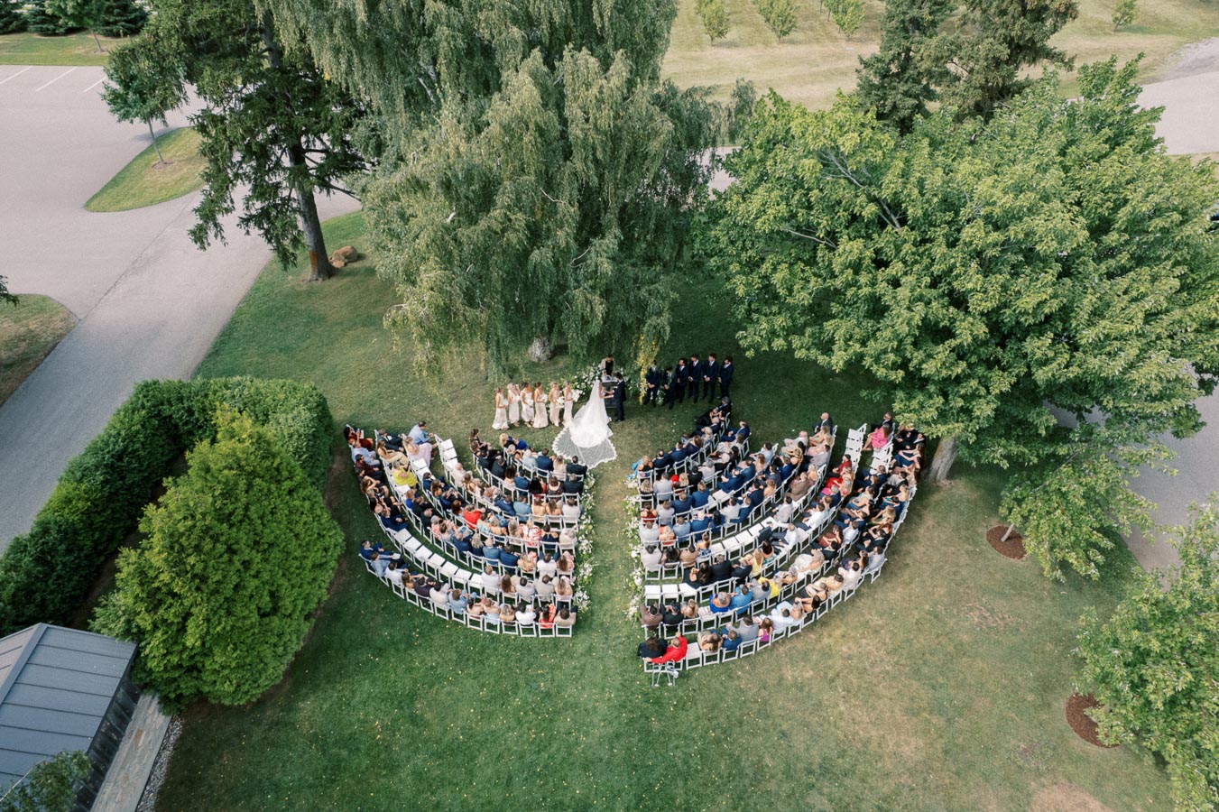 Aerial view of an outdoor wedding ceremony with guests seated in rows forming a semi-circle under lush green trees.